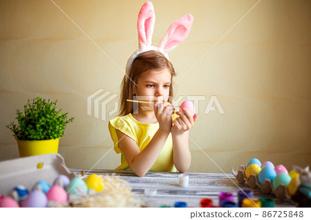 Cute girl in bunnys ears paints Easter eggs at home while sitting at the table. Preparing for Easter Cute girl in bunnys ears paints Easter eggs at home while sitting at the table. Preparing for Easter 86725848
