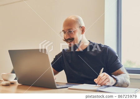 Young caucasian freelancer works with laptop at his desk in office while doing something notes Young caucasian freelancer works with laptop at his desk in office while doing something notes 86725979