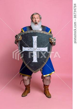 Shock. Portrait of senior grey bearded man, brave medieval warrior or knight in armored clothes with sword isolated on pink background. Comparison of eras, history, festival Shock. Portrait of senior grey bearded man, brave medieval warrior or knight in armored clothes with sword isolated on pink background. Comparison of eras, history, festival 86734784