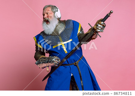 Portrait of senior grey bearded man, brave medieval warrior or knight in armored clothes with sword isolated on pink background. Comparison of eras, history, festival 86734811