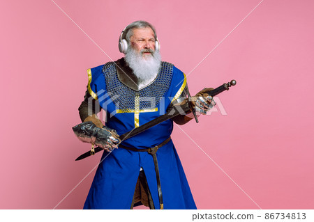 Portrait of senior grey bearded man, brave medieval warrior or knight in armored clothes with sword isolated on pink background. Comparison of eras, history, festival Portrait of senior grey bearded man, brave medieval warrior or knight in armored clothes with sword isolated on pink background. Comparison of eras, history, festival 86734813