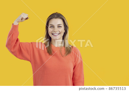 Studio shot of happy confident strong cheerful good looking young Caucasian woman standing on yellow background, smiling at camera and flexing one arm, demonstrating her superpower. Girl power concept Studio shot of happy confident strong cheerful good looking young Caucasian woman standing on yellow background, smiling at camera and flexing one arm, demonstrating her superpower. Girl power concept 86735513