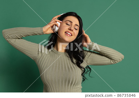 Happy young beautiful girl with long glossy dark hair listening to music isolated on green background. Concept of emotions, facial expression Happy young beautiful girl with long glossy dark hair listening to music isolated on green background. Concept of emotions, facial expression 86735846