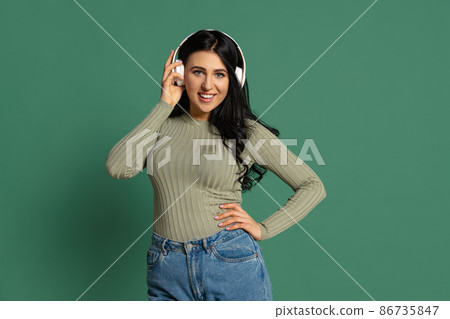 Happy young beautiful girl with long glossy dark hair listening to music isolated on green background. Concept of emotions, facial expression 86735847