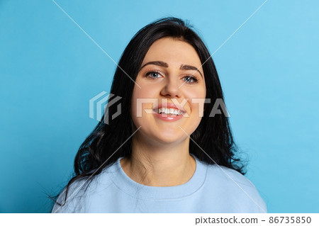 Portrait of young beautiful girl with long glossy dark hair posing isolated on blue background. Concept of emotions, facial expression Portrait of young beautiful girl with long glossy dark hair posing isolated on blue background. Concept of emotions, facial expression 86735850