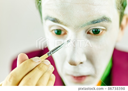 Close up of clown cosplayer. Mim is preparing makeup for performance. Man with green hair in burgundy suit. Theatre atmosphere. Male actor is covering his face by white greasepaint using brush. 86738398