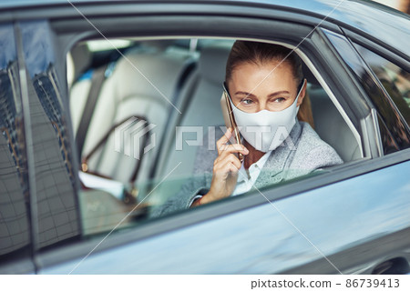 Covid 19 or coronavirus outbreak, caucasian businesswoman wearing medical protective mask talking on smartphone while sitting on back seat in the car, looking out of a window Covid 19 or coronavirus outbreak, caucasian businesswoman wearing medical protective mask talking on smartphone while sitting on back seat in the car, looking out of a window 86739413