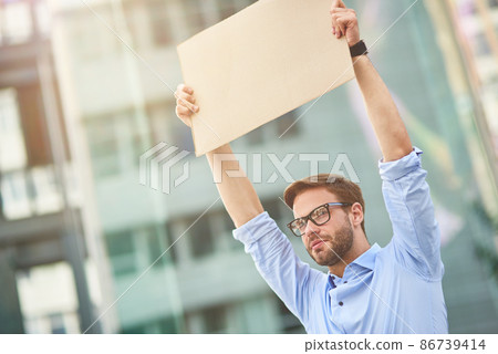 Protesting outdoors. Portrait of a young male activist wearing blue shirt and eyeglasses holding empty sign board over the head while standing outdoors Protesting outdoors. Portrait of a young male activist wearing blue shirt and eyeglasses holding empty sign board over the head while standing outdoors 86739414