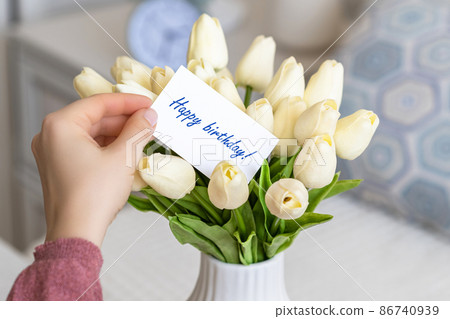 Woman pulling greeting card with Happy birthday words from bouquet of white tulips flowers in a white vase on the table. Woman pulling greeting card with Happy birthday words from bouquet of white tulips flowers in a white vase on the table. 86740939