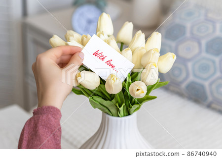 Woman pulling greeting card with love words (text) from bouquet of white tulips flowers in a white vase on the table. Woman pulling greeting card with love words (text) from bouquet of white tulips flowers in a white vase on the table. 86740940