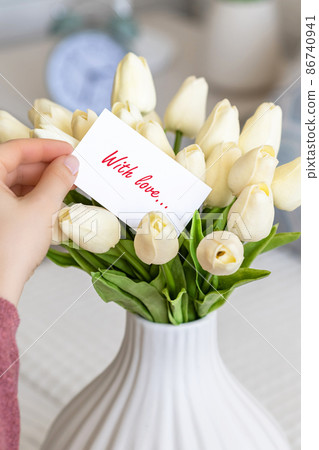 Woman pulling greeting card with love words (text) from bouquet of white tulips flowers in a white vase on the table. 86740941