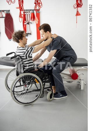 Rehabilitation specialist helps man to get up from a wheelchair at clinic Rehabilitation specialist helps man to get up from a wheelchair at clinic 86741382