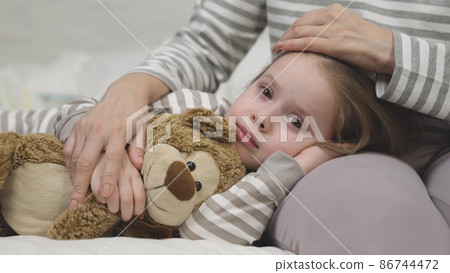 Small girl hugs teddy bear lying on mother lap before sleep Small girl hugs teddy bear lying on mother lap before sleep 86744472