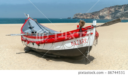 Nazare, Portugal - April 2018: Colorful traditional old wooden fishing boat on the beach of fishing village of Nazare. The high cliff of Nazare Sitio and its lighthouse fortress on background. Nazare, Portugal - April 2018: Colorful traditional old wooden fishing boat on the beach of fishing village of Nazare. The high cliff of Nazare Sitio and its lighthouse fortress on background. 86744531
