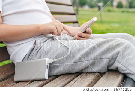 Boy using phone on the bench while charging from the power bank. 86745296
