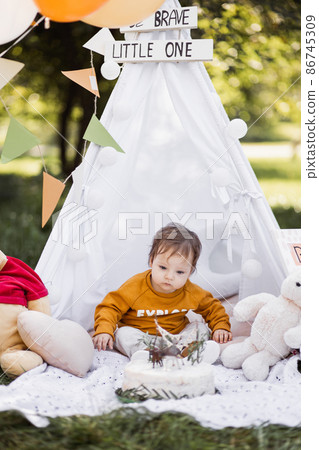 Cute baby boy sitting in wigwam near his birthday cake Cute baby boy sitting in wigwam near his birthday cake 86745309