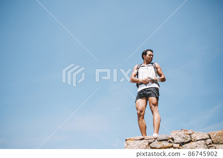 Active young man with backpack standing on cliff Active young man with backpack standing on cliff 86745902