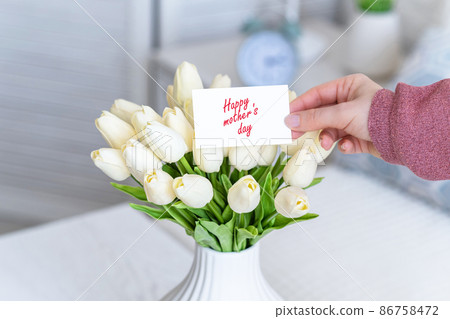 Woman pulling greeting card with Happy mother's day words from bouquet of white tulips flowers in a white vase on the table. 86758472