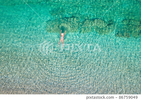Top, aerial view. Young beautiful woman in a hat and white bikini swimming in sea water on the sand beach. Drone, copter photo. Summer vacation. View from above. Top, aerial view. Young beautiful woman in a hat and white bikini swimming in sea water on the sand beach. Drone, copter photo. Summer vacation. View from above. 86759049