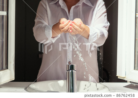 Water drops pouring down through female hands during morning washing. Female standing near open window of bathroom at sunny summer 86762458