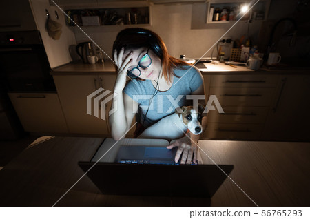 Tired woman in headphones and glasses studying at a laptop. A female student prepares for an exam in the kitchen late in the evening. Girl with a puppy on her knees. Tired woman in headphones and glasses studying at a laptop. A female student prepares for an exam in the kitchen late in the evening. Girl with a puppy on her knees. 86765293