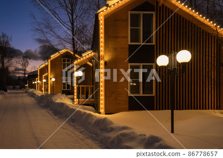 A snow-covered street of houses in the style of a barnhouse. The houses are decorated with Christmas garlands. Warm yellow backlight 86778657