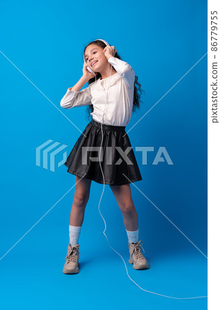 Schoolgirl with headphones listening to music against blue background Schoolgirl with headphones listening to music against blue background 86779755