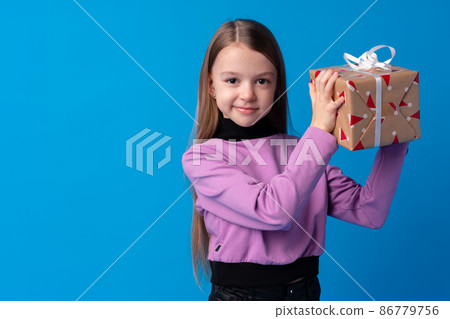 Happy little girl with long hair holding a gift box on a blue background Happy little girl with long hair holding a gift box on a blue background 86779756