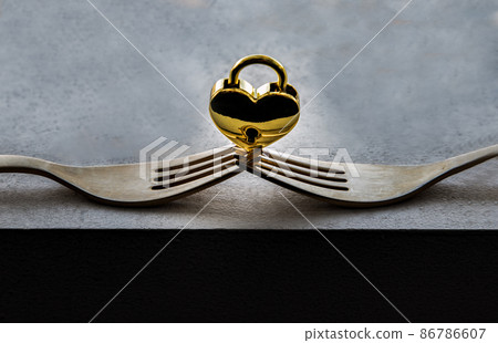 Golden heart shaped love padlock balanced on a composition of two intertwined gold forks. 86786607