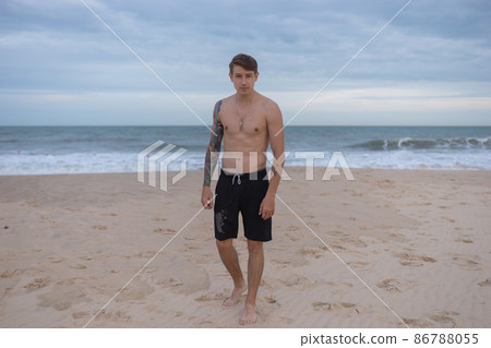 Full height portrait of handsome fit bare-chested young man standing on the beach. guy with tattoo walking by the sea. High quality photo Full height portrait of handsome fit bare-chested young man standing on the beach. guy with tattoo walking by the sea. High quality photo 86788055