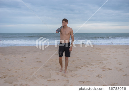 Full height portrait of handsome fit bare-chested young man standing on the beach. guy with tattoo walking by the sea. High quality photo 86788056