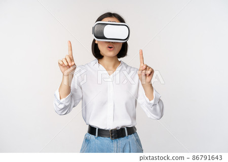 Image of asian woman in vr glasses pointing fingers up, looking amazed and surprised. Girl using virtual reality headset, concept of future and communication, white background 86791643