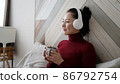 Mixed race woman holding cup of tea or coffee drinking warm beverage at home. Young asian girl in headphones listening chill music with morning drink looking through window enjoying and dreaming 86792754