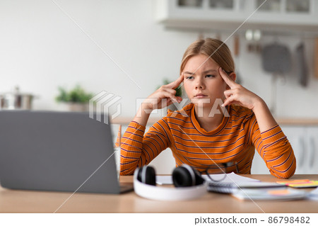 Tired sad concentrated european teen girl presses fingers to head, watches video lesson on laptop Tired sad concentrated european teen girl presses fingers to head, watches video lesson on laptop 86798482