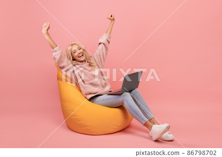 Overjoyed woman in casual sitting in beanbag chair with laptop and raising hands up, got dream job or great news 86798702