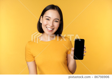 Beautiful happy asian girl showing mobile phone screen, application on smartphone gadget, standing over yellow background Beautiful happy asian girl showing mobile phone screen, application on smartphone gadget, standing over yellow background 86798906