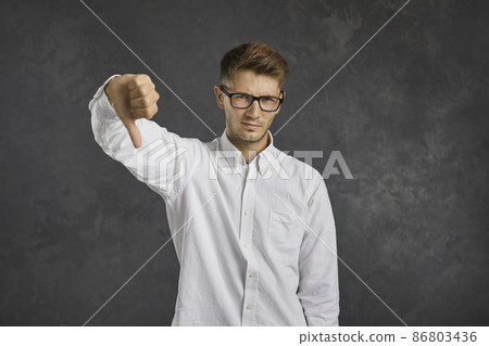 Dissatisfied and frustrated young man showing thumb down standing on gray background. Caucasian man in glasses and shirt gives a negative answer or feedback. Concept of dislike and disapproval. Dissatisfied and frustrated young man showing thumb down standing on gray background. Caucasian man in glasses and shirt gives a negative answer or feedback. Concept of dislike and disapproval. 86803436