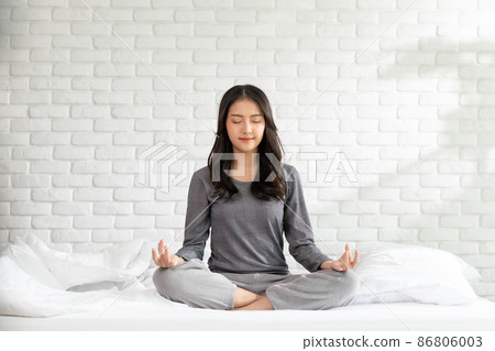 Calm of wellness Asian young woman sit on bed breathing with yoga lotus pose,Yoga meditation of young healthy woman relax and comfortable at white cozy home,Yoga Exercise for Wellness Concept 86806003