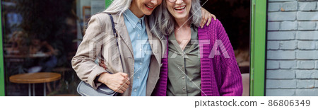 Pretty senior Asian lady hugs friend in purple jacket standing near cafe on street Pretty senior Asian lady hugs friend in purple jacket standing near cafe on street 86806349