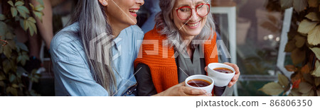 Smiling mature Asian lady with friend clink cups of coffee sitting near cafe Smiling mature Asian lady with friend clink cups of coffee sitting near cafe 86806350