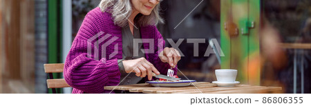 Pretty senior lady in purple knitted jacket eats delicious dessert at table on outdoors cafe terrace Pretty senior lady in purple knitted jacket eats delicious dessert at table on outdoors cafe terrace 86806355