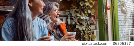 Positive mature women companions with cups sit near window of cafe Positive mature women companions with cups sit near window of cafe 86806357