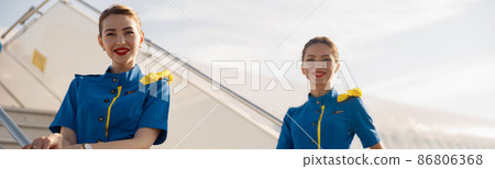 Two beautiful air stewardesses in blue uniform smiling at camera, standing on airstair and welcoming passengers 86806368
