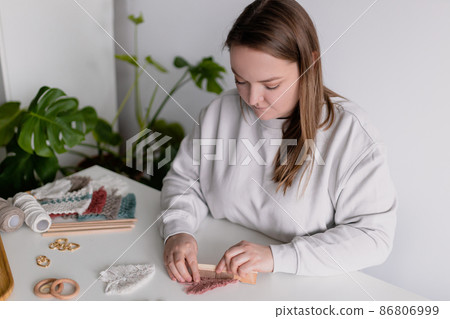 Woman working out last details of an almost-finished macrame piece, combing ropes inside a home room. Close up. Natural cotton threads and wooden beads. Female hobby. Wall hanging decor. Woman working out last details of an almost-finished macrame piece, combing ropes inside a home room. Close up. Natural cotton threads and wooden beads. Female hobby. Wall hanging decor. 86806999
