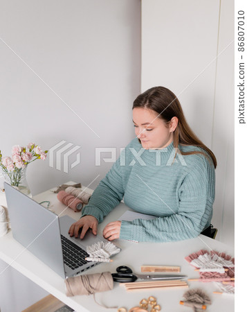 Young stylish woman using laptop at home, looking at screen, chatting, reading or writing email. Female hobby. Working on research project online concept. Handmade macrame shop 86807010