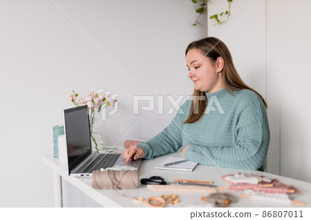 Young stylish woman using laptop at home, looking at screen, chatting, reading or writing email. Female hobby. Working on research project online concept. Handmade macrame shop 86807011