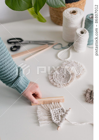 Woman working out last details of an almost-finished macrame piece, combing ropes inside a home room. Close up. Natural cotton threads and wooden beads. Female hobby. Wall hanging decor. 86807012
