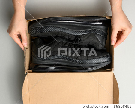 female hands are holding a brown cardboard box with black sports sneakers on a gray table, inspecting an online order, top view 86808495