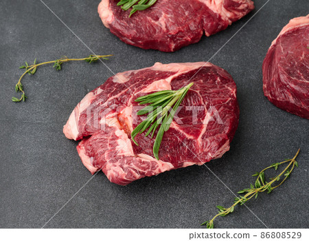 raw piece of beef ribeye with rosemary, thyme on a black table, top view 86808529