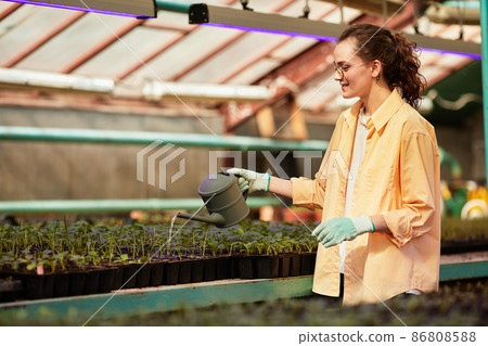 Young happy woman in gloves, shirt and eyeglasses watering sprouts Young happy woman in gloves, shirt and eyeglasses watering sprouts 86808588